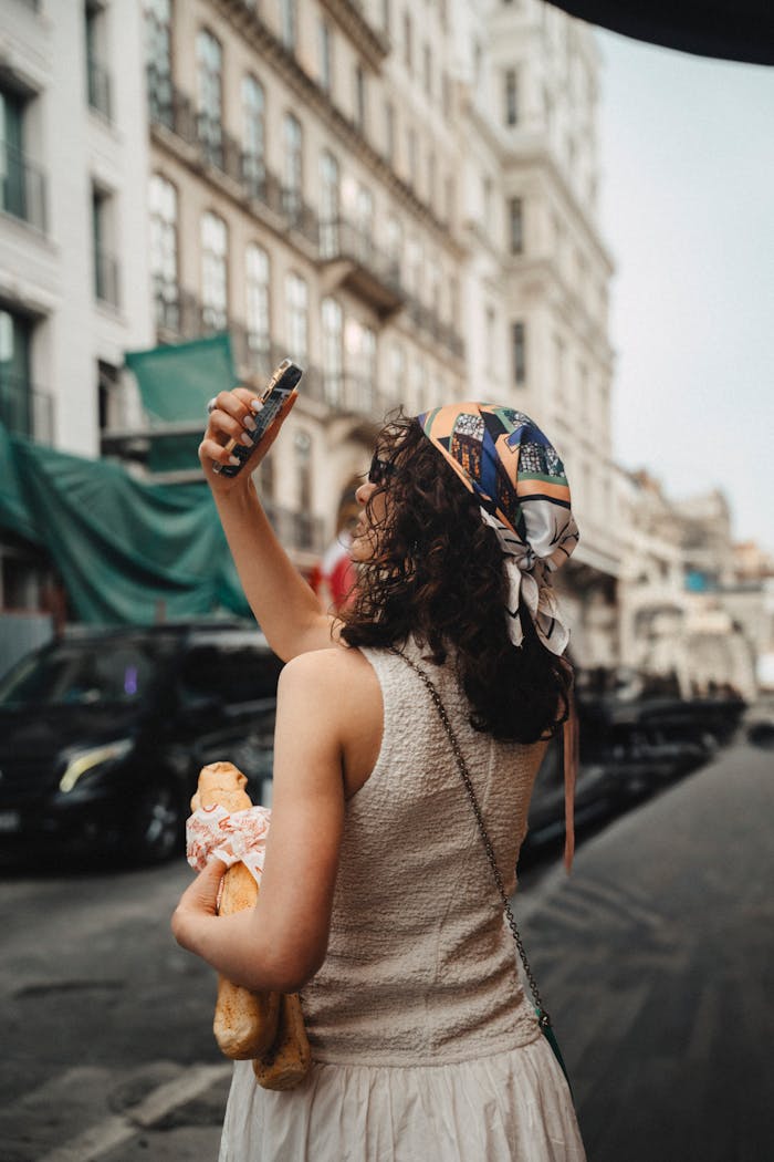 Young woman with headscarf takes a selfie in a European city, holding a baguette.
